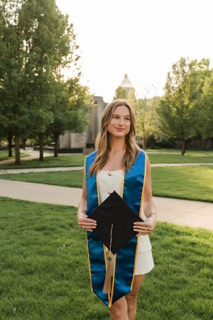Graduate in blue and white stole holds graduation cap on university lawn with building in background.