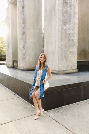 Graduate in blue stole poses between white columns in sunny outdoor setting.