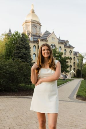 Graduate in white dress poses in front of Notre Dame's golden dome building.