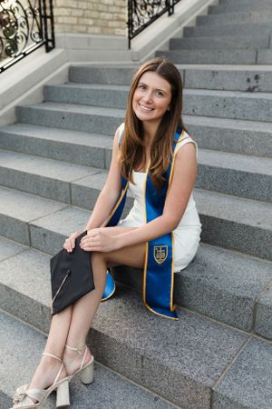 Graduate in white dress and blue sash sits on stone steps during commencement at Notre Dame.