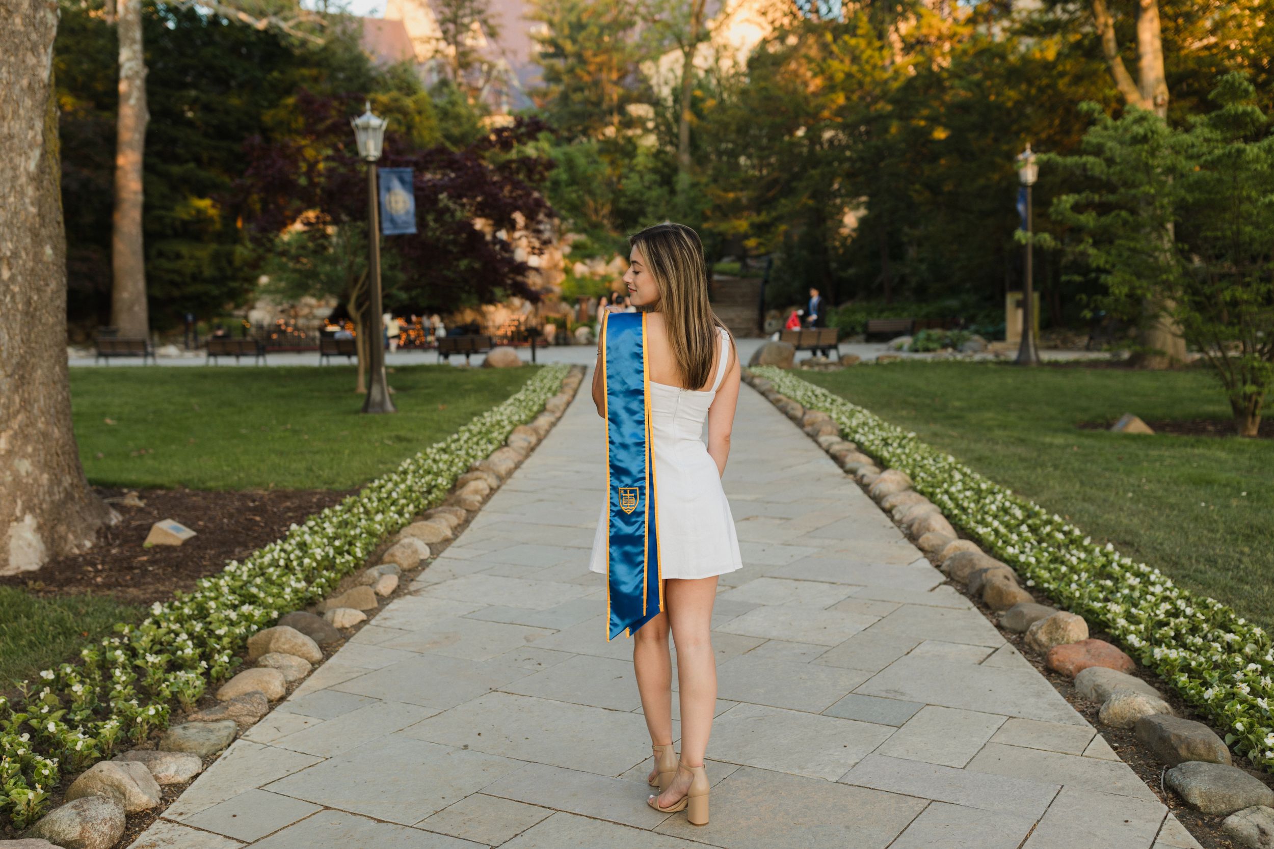 Person in a white dress holding blue graduation stole while walking on a garden path.