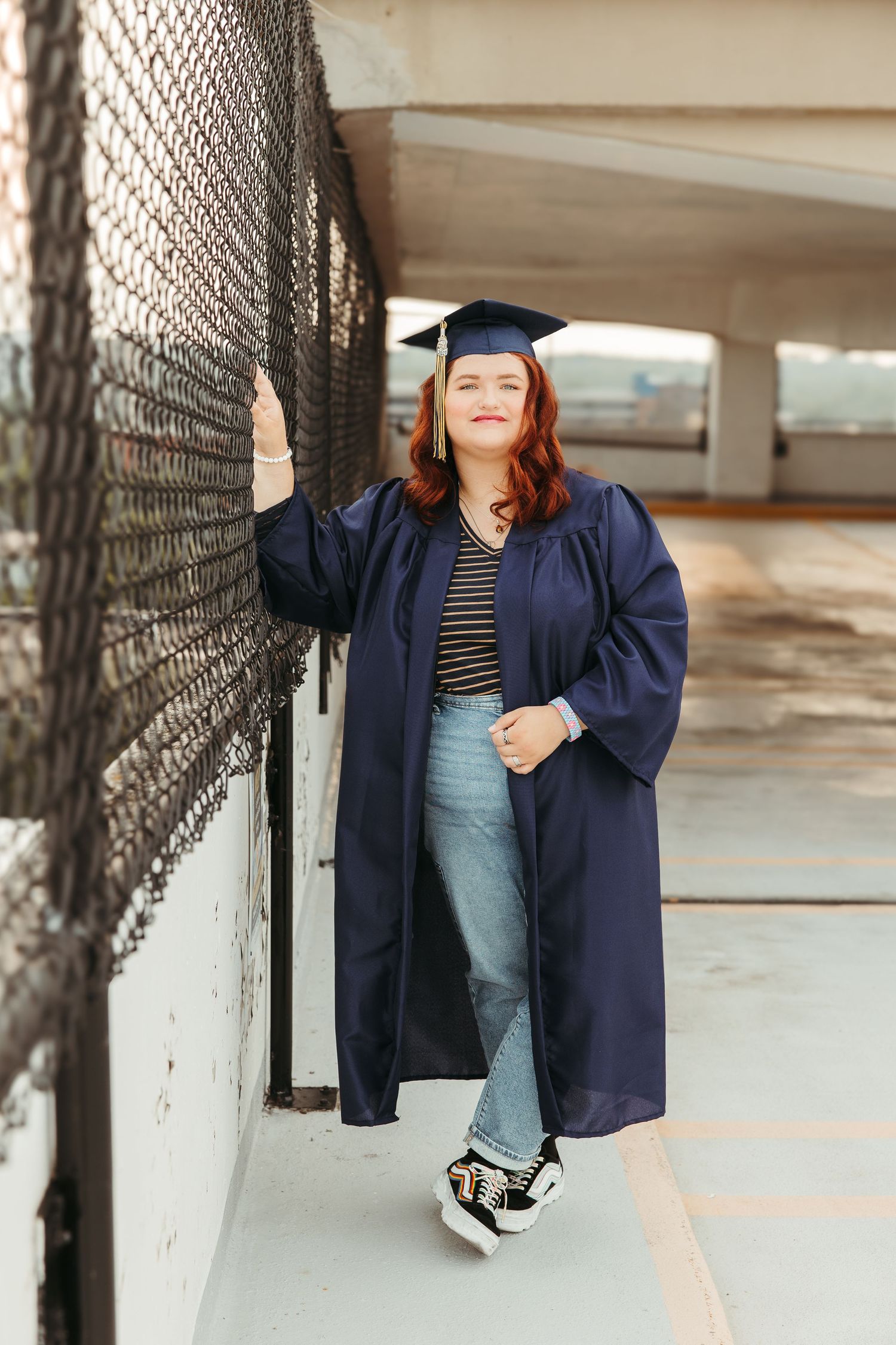 Graduate wearing navy blue cap and gown poses in a parking garage while leaning against chain link fence.