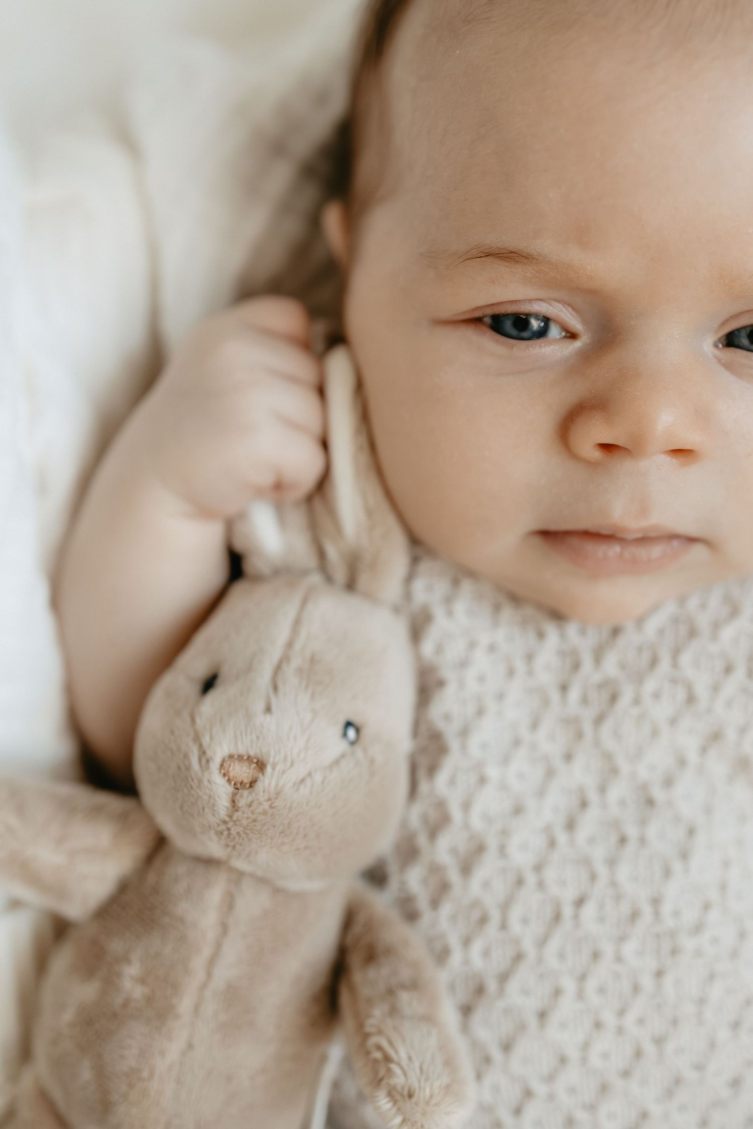 Suloinen vauva makaa valkoisella teksturoidulla vuodevaatteella pehmeän pehmolelupupun kanssa. Vastasyntyneen kuvaus.