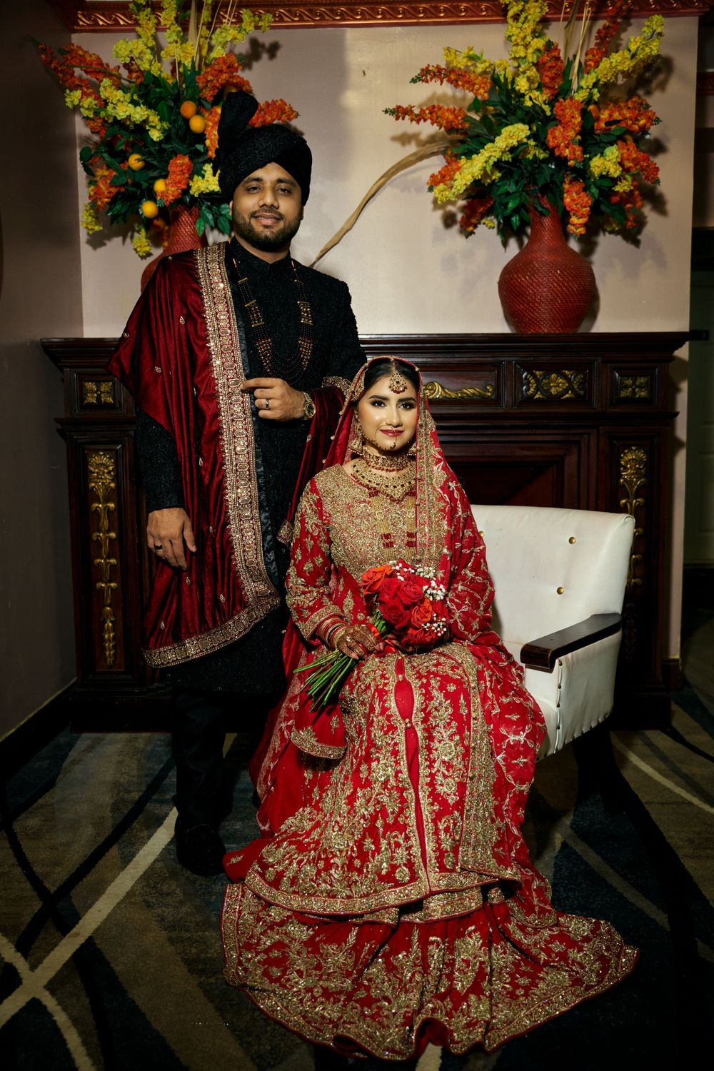 South Asian bride and groom in traditional red and gold wedding attire pose with floral arrangements.