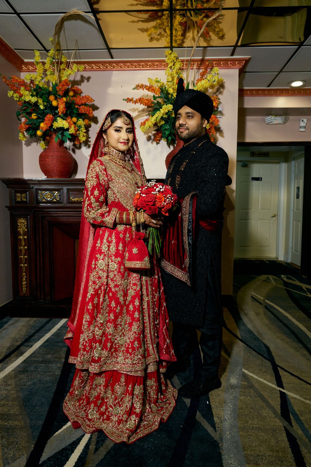 Couple in traditional South Asian wedding attire pose with red bouquet against floral decor.