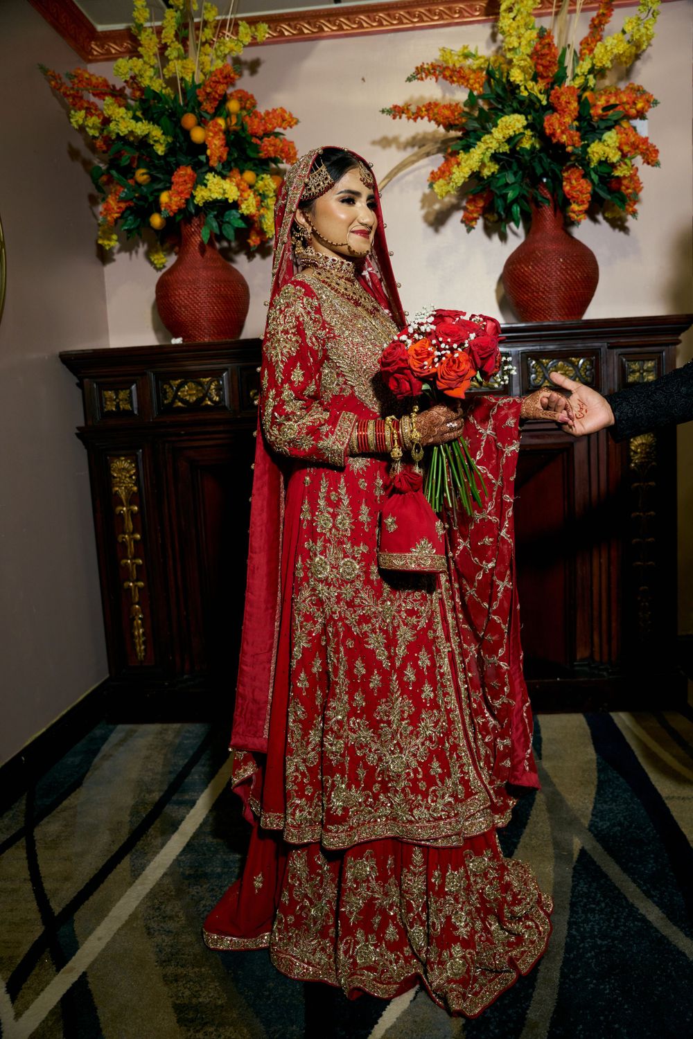 South Asian bride in red embroidered lehenga holds bouquet against floral backdrop.