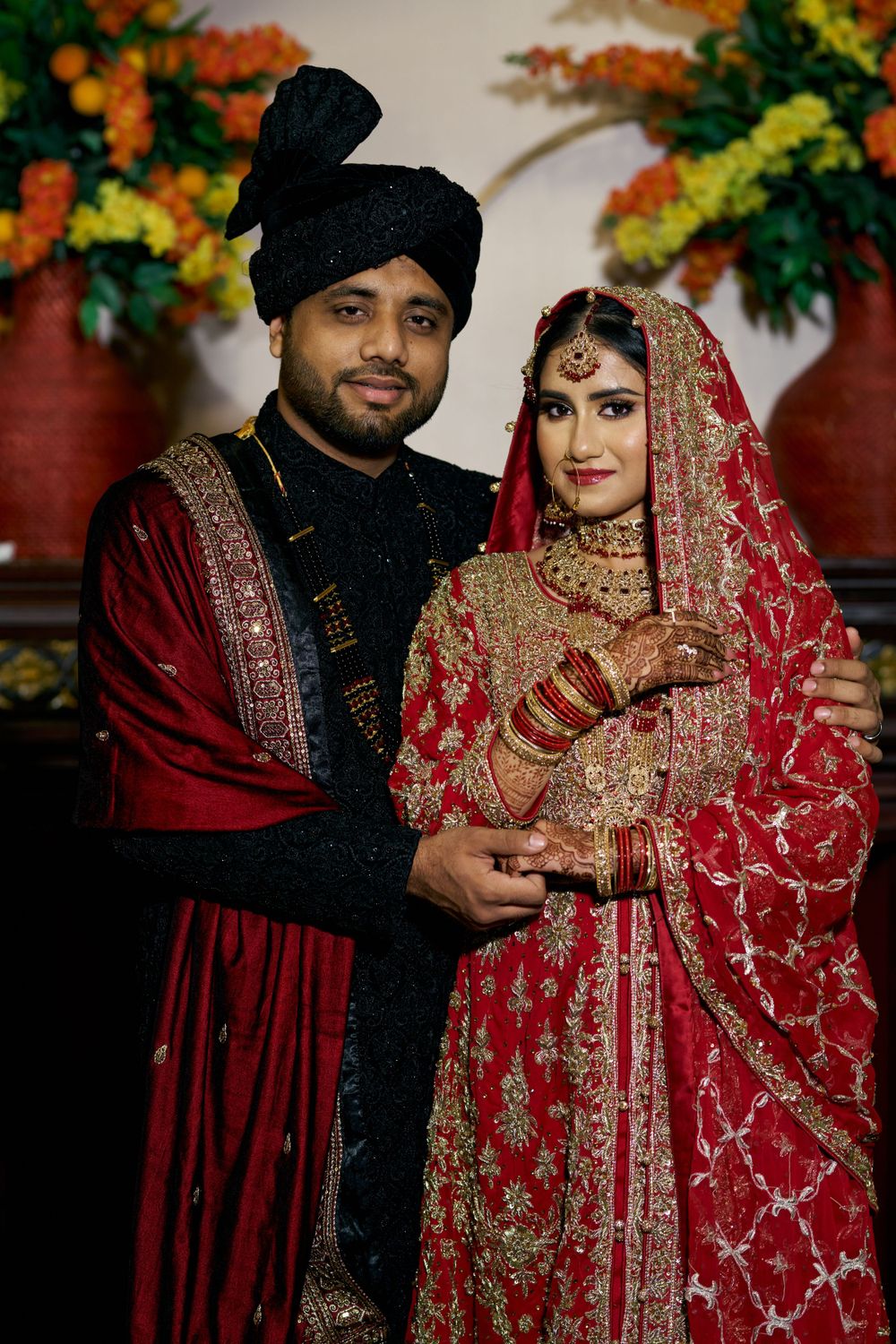 South Asian wedding couple in elegant red and black traditional attire with floral decorations.