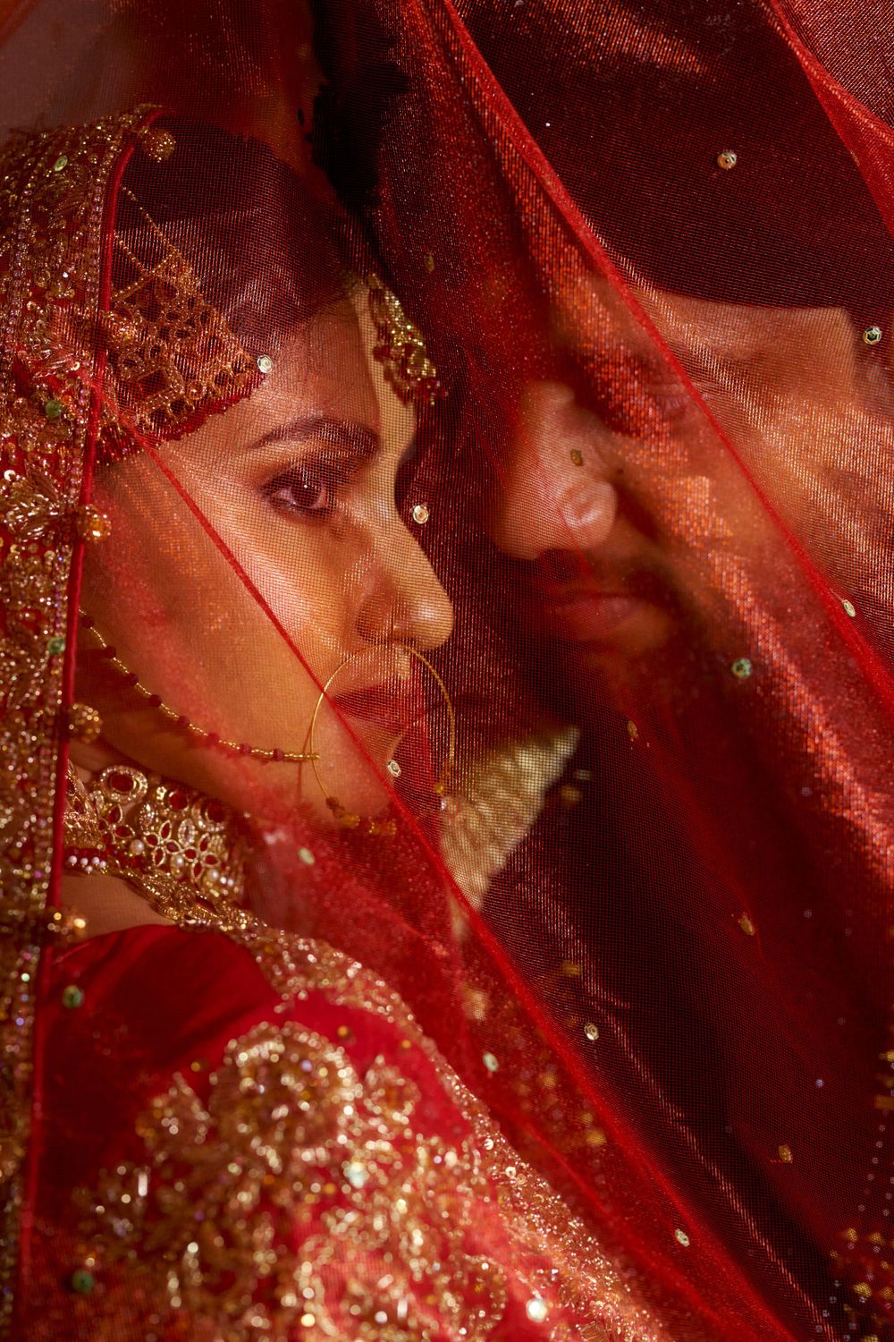 Artistic portrait through sheer red bridal veil with sparkling golden details.
