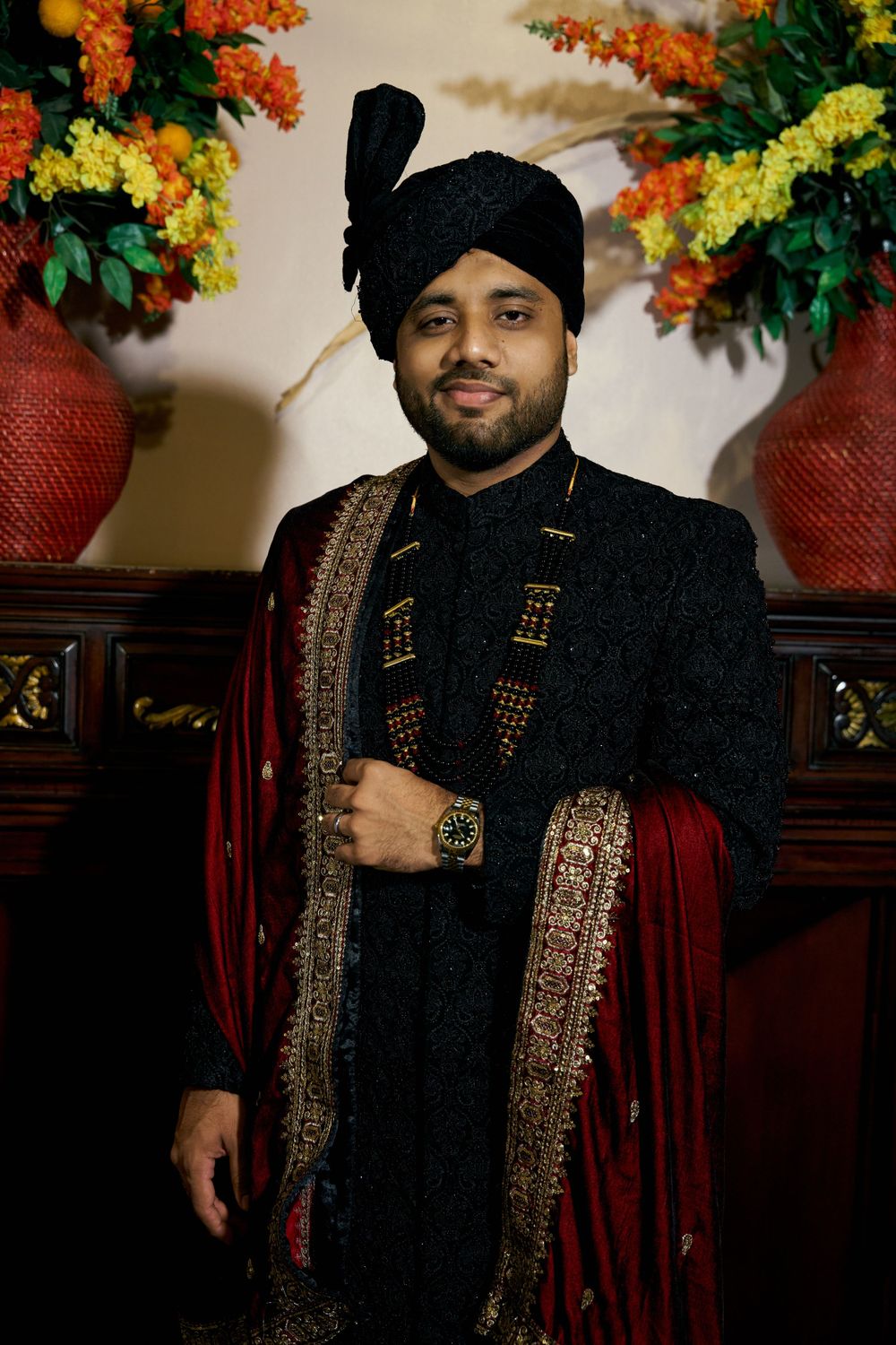 Groom in traditional black Punjabi wedding attire with ornate embroidery against floral backdrop.