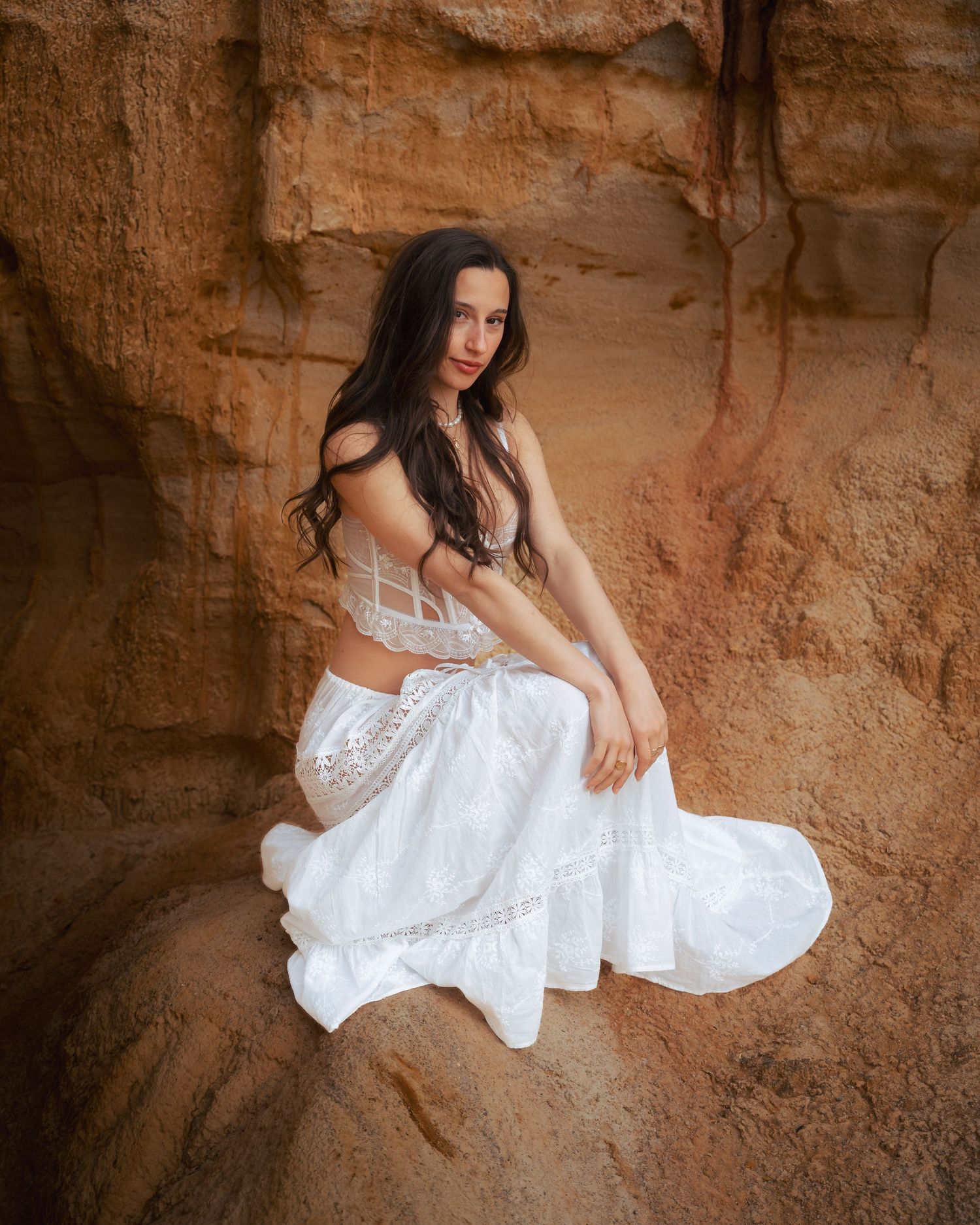 Woman in white dress poses against textured orange canyon wall in artistic portrait.