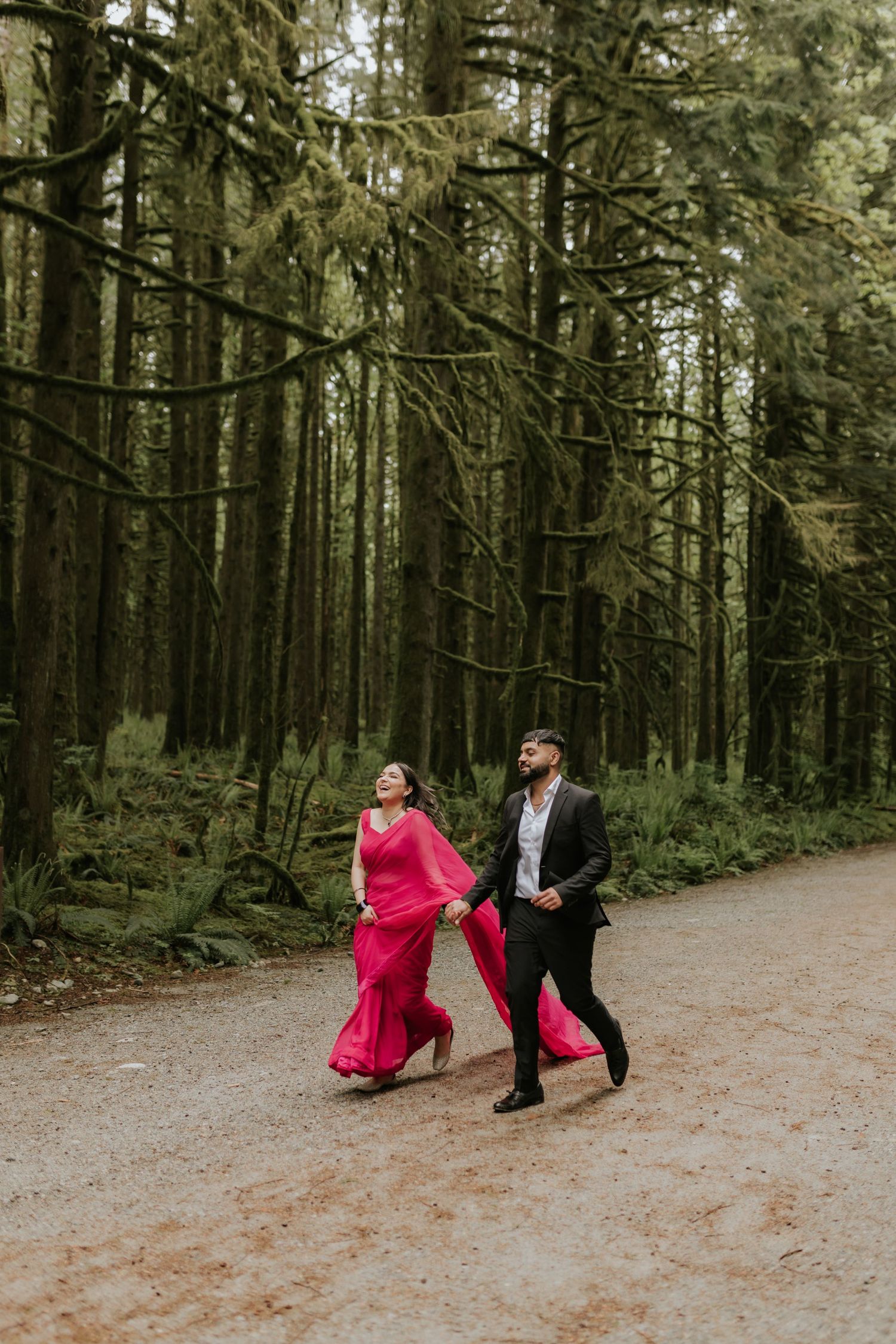 Couple in formal attire walking together on a forest path surrounded by tall evergreen trees.