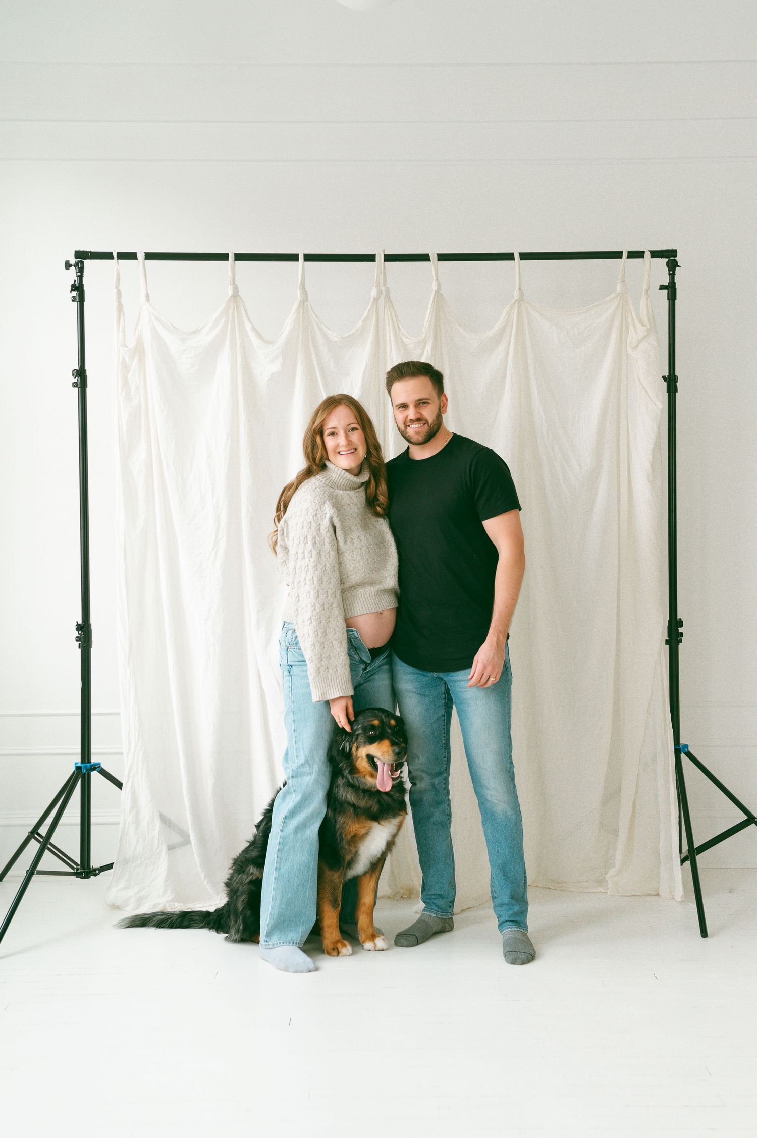 A couple stands with their German Shepherd mix dog against a white backdrop in a photography studio setting.