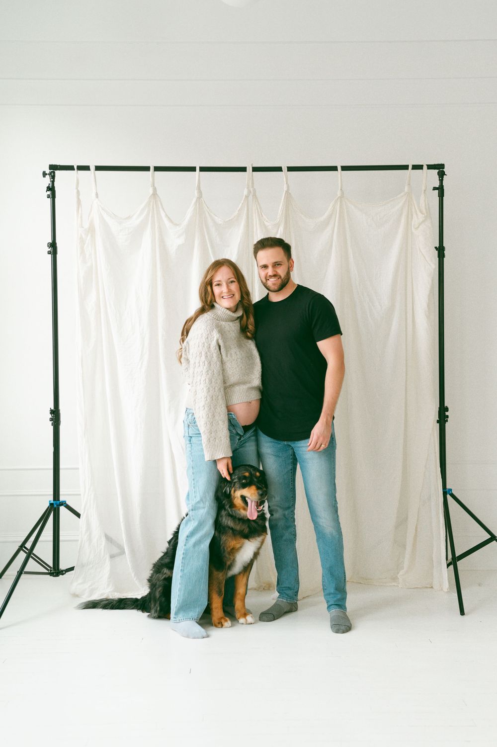 A couple stands with their German Shepherd mix dog against a white backdrop in a photography studio setting.