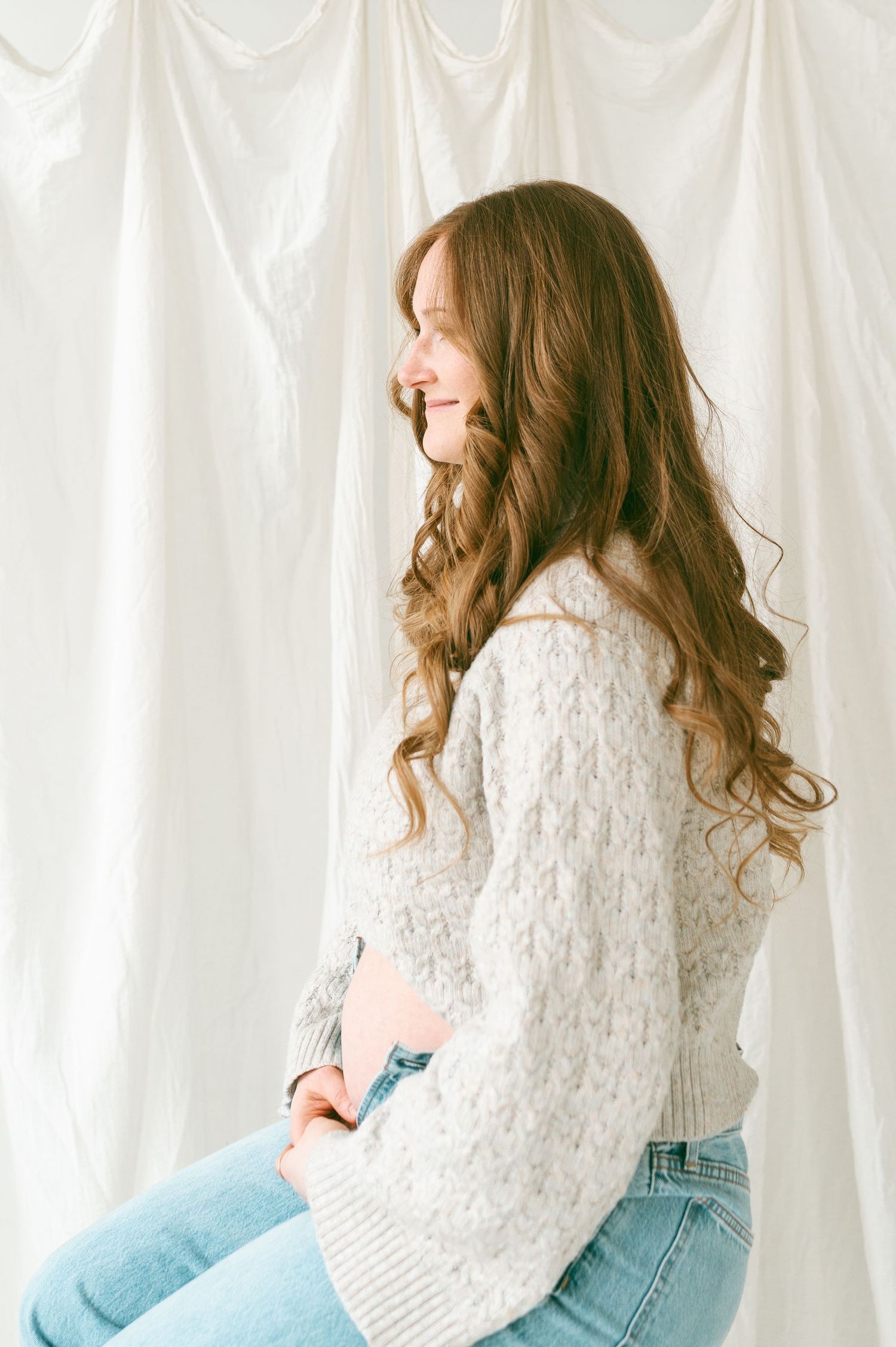 Side profile of someone in a cozy white knit sweater and light jeans sitting against a white curtain backdrop.