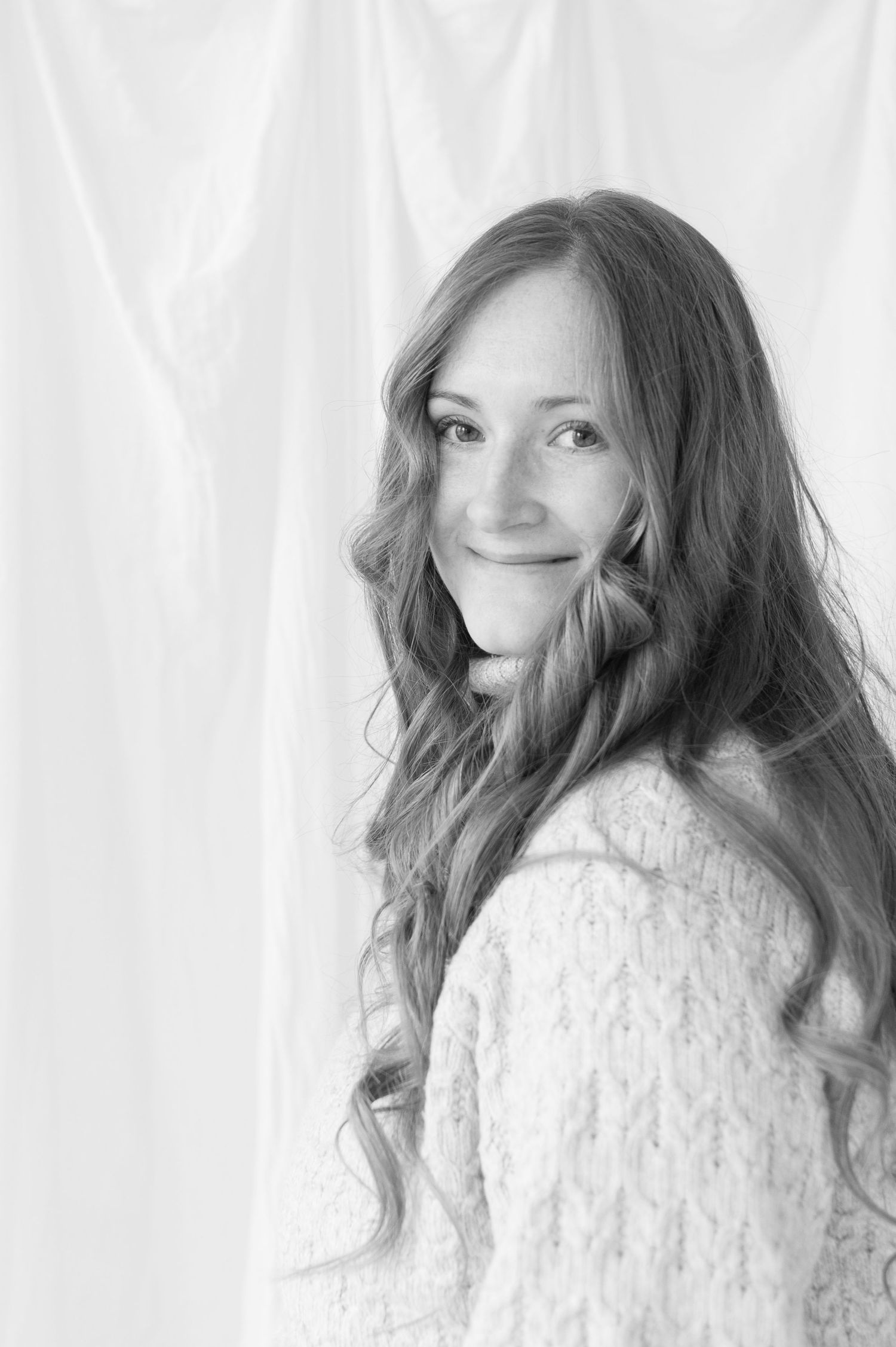 A black and white portrait of someone with long wavy hair wearing a light-colored knit sweater against a white backdrop.