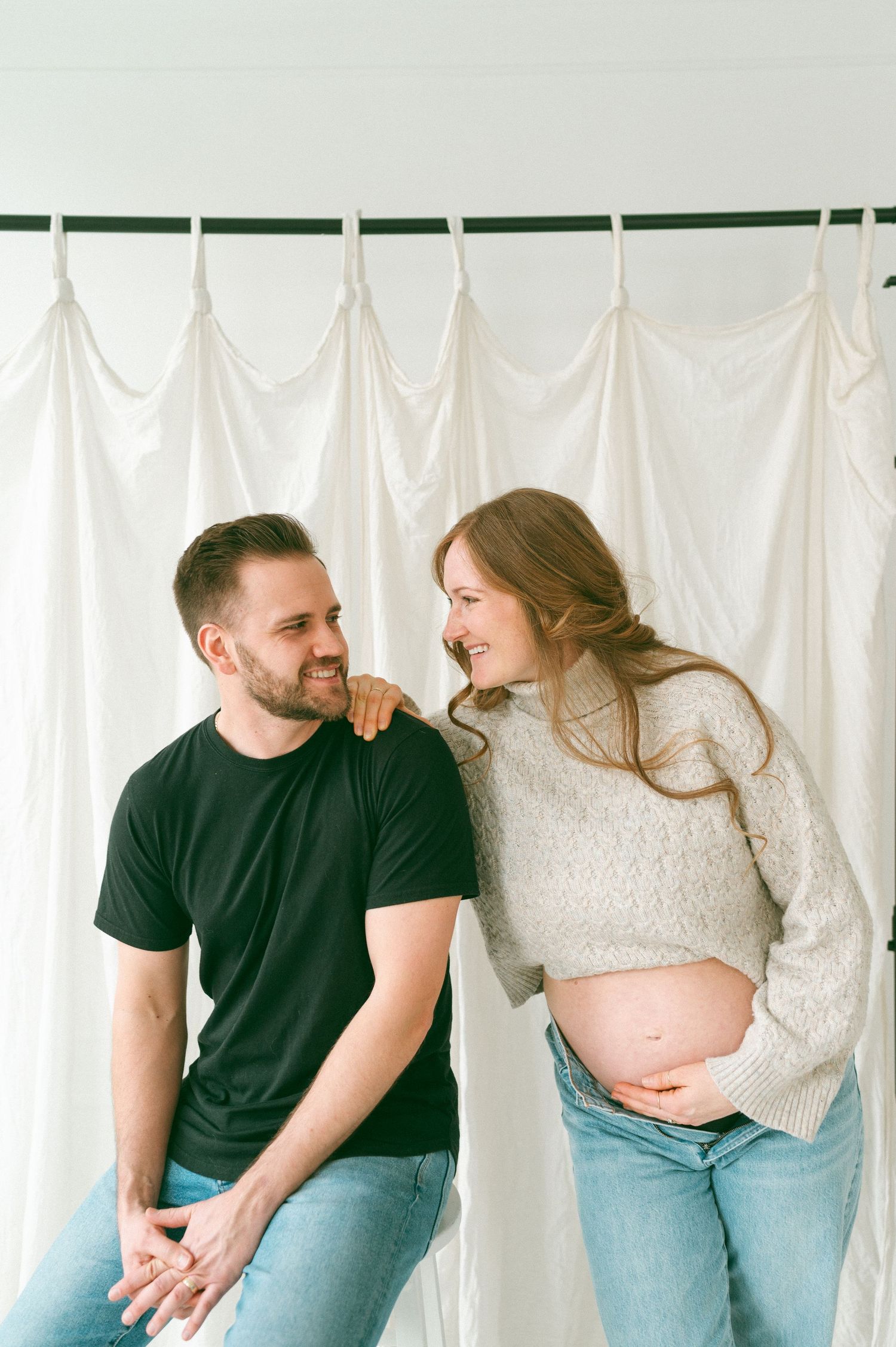 Expecting couple share loving moment against white curtain backdrop while showcasing a baby bump in casual attire.