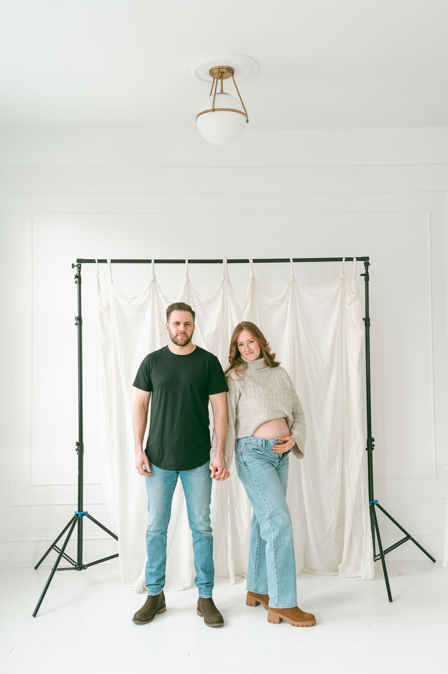 Two people stand against a white backdrop in casual attire in a minimalist photography studio setting.