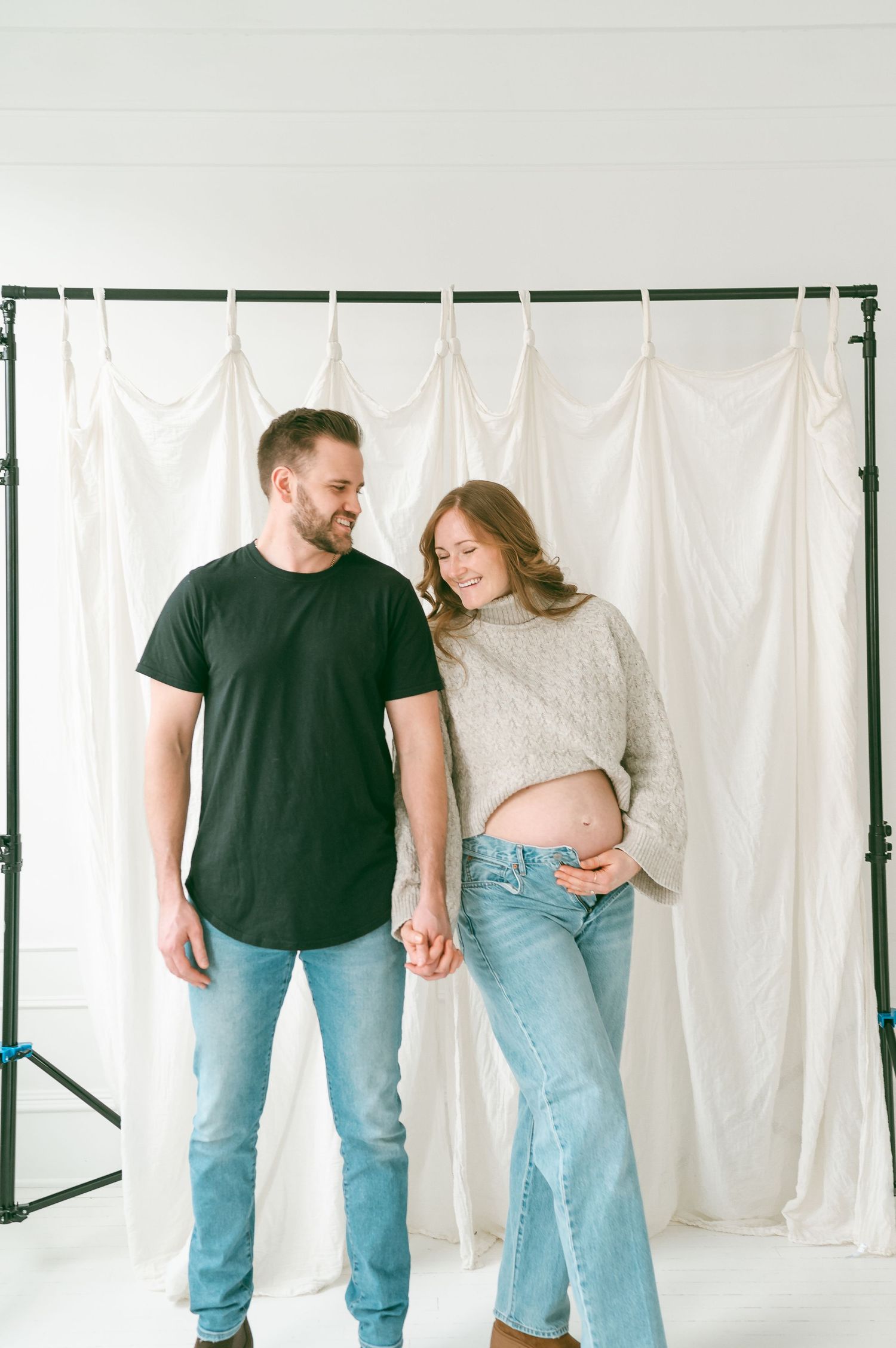 A couple in casual attire shares a joyful moment together against a white backdrop during a photo session.