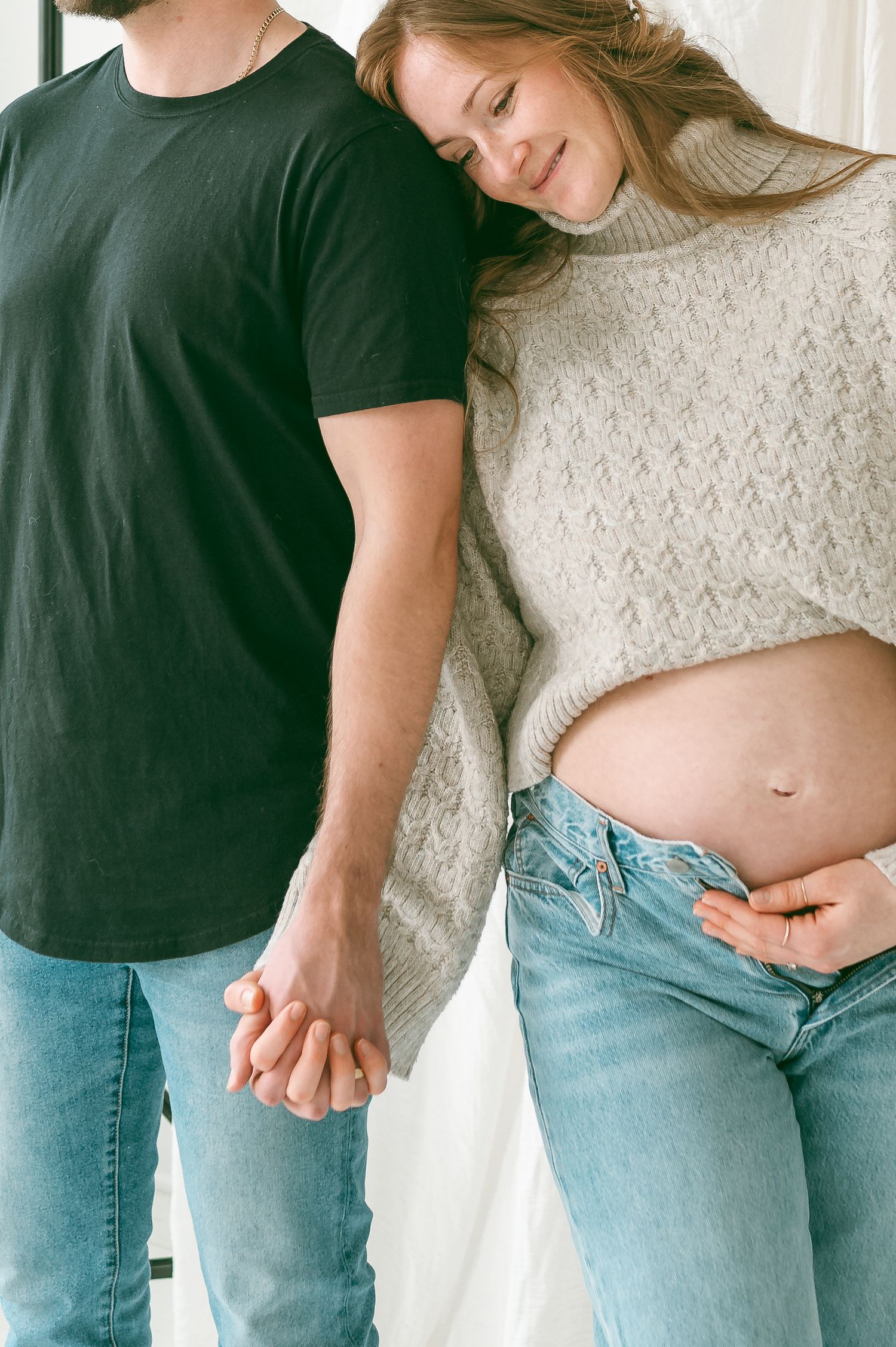 A couple in casual wear holding hands with woman displaying her pregnant belly in a knit sweater and jeans.