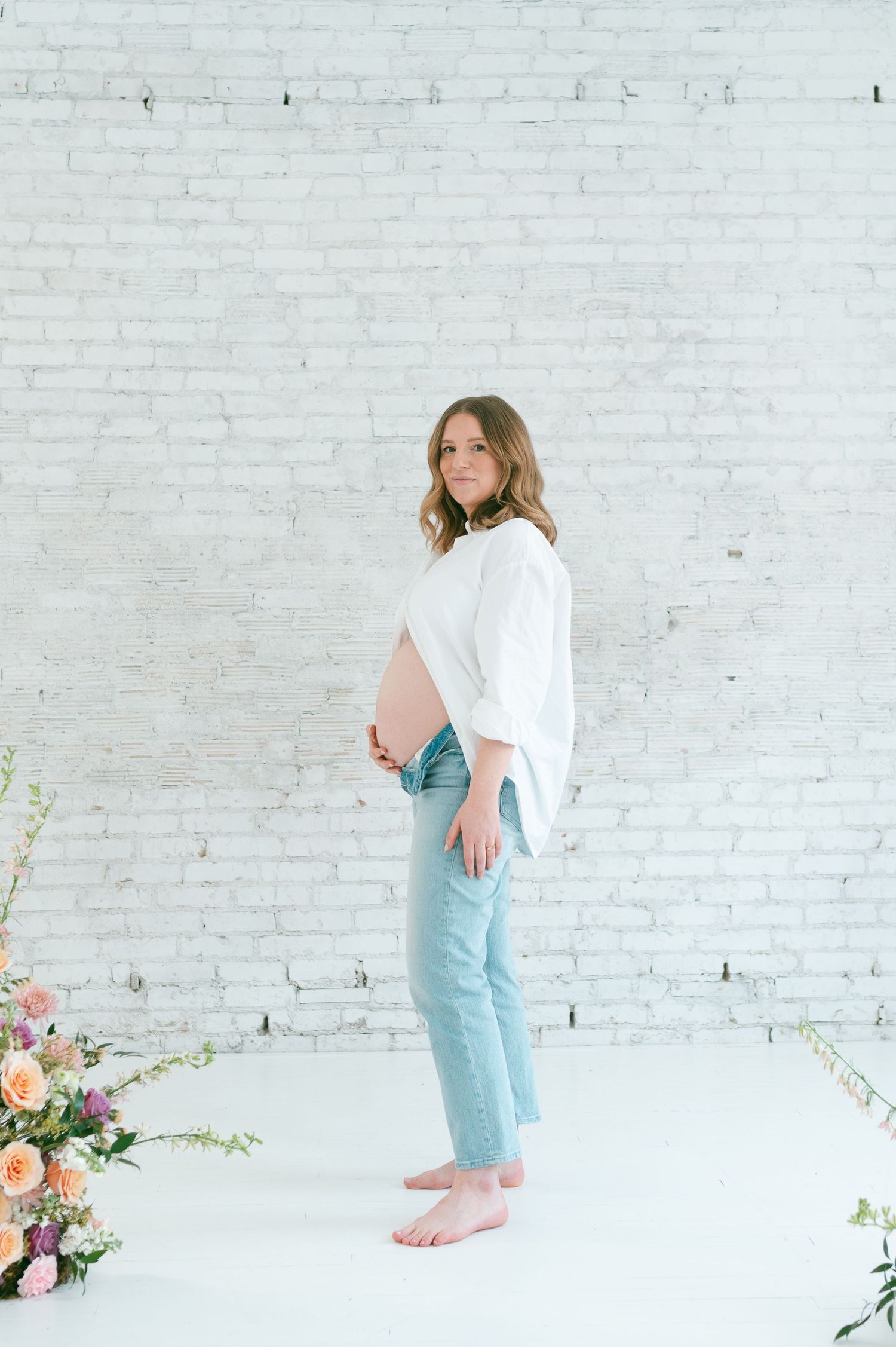 Maternity portrait against white brick wall wearing casual white shirt and jeans.