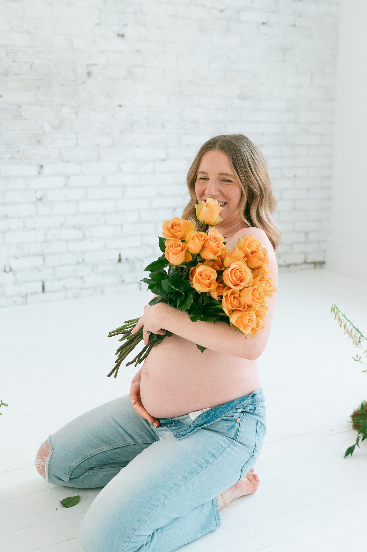 Joyful maternity photo with subject holding bouquet of orange roses against white backdrop.