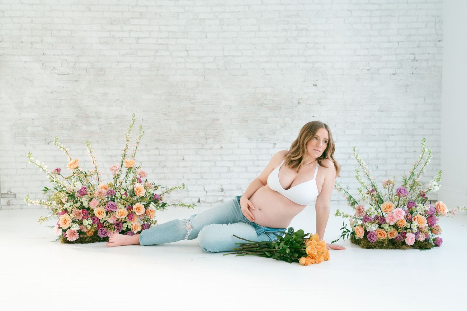 Maternity portrait with floral arrangements and soft lighting against white brick wall.