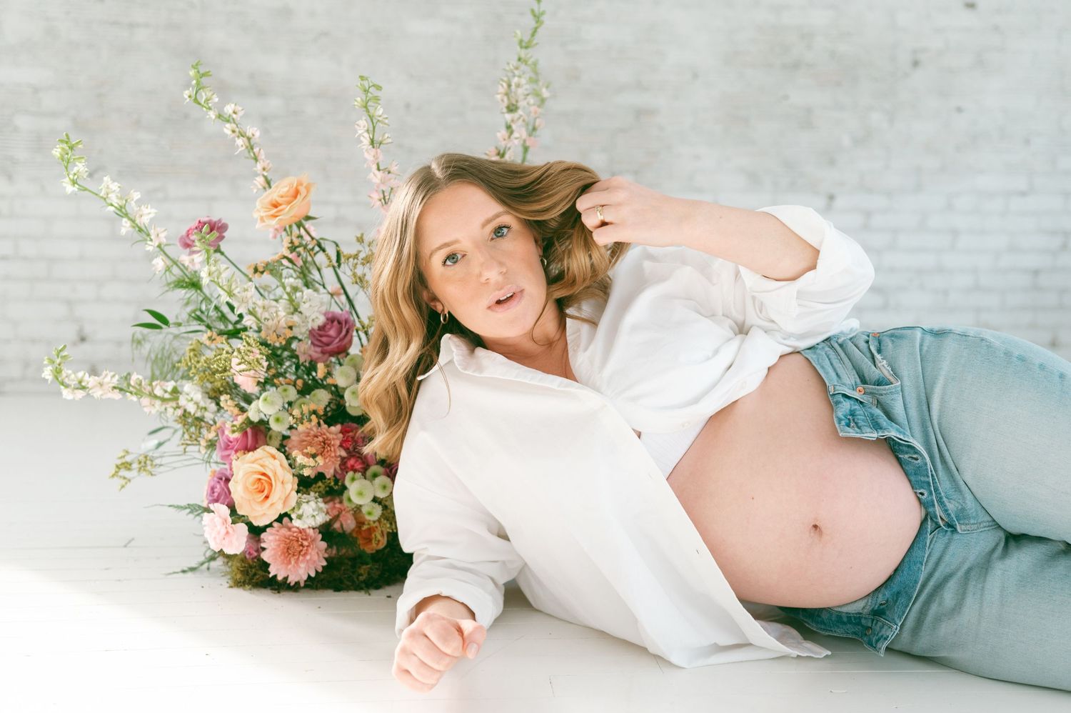 Expectant mother lying on floor with floral arrangement in white studio setting.