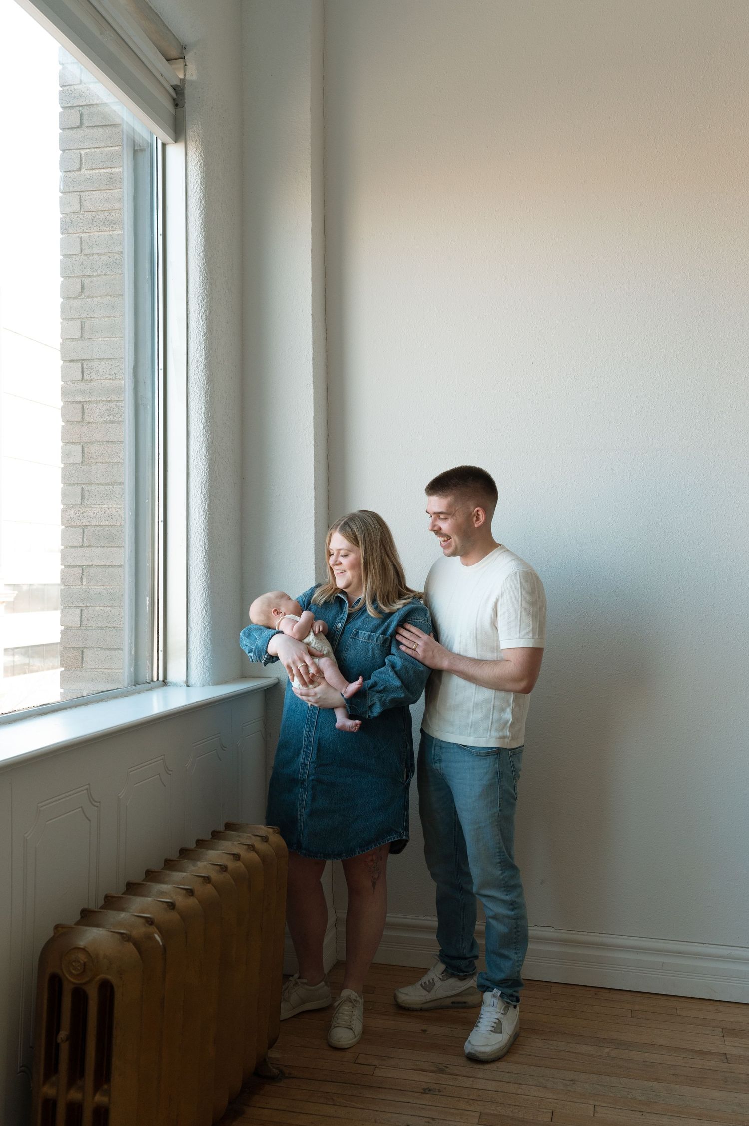 A couple stands by a sunny window holding their newborn baby in a bright minimalist room with hardwood floors and a radiator.