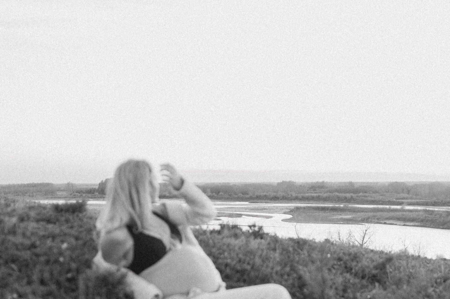 Black and white photo of a person lounging on a hillside overlooking a scenic waterway.
