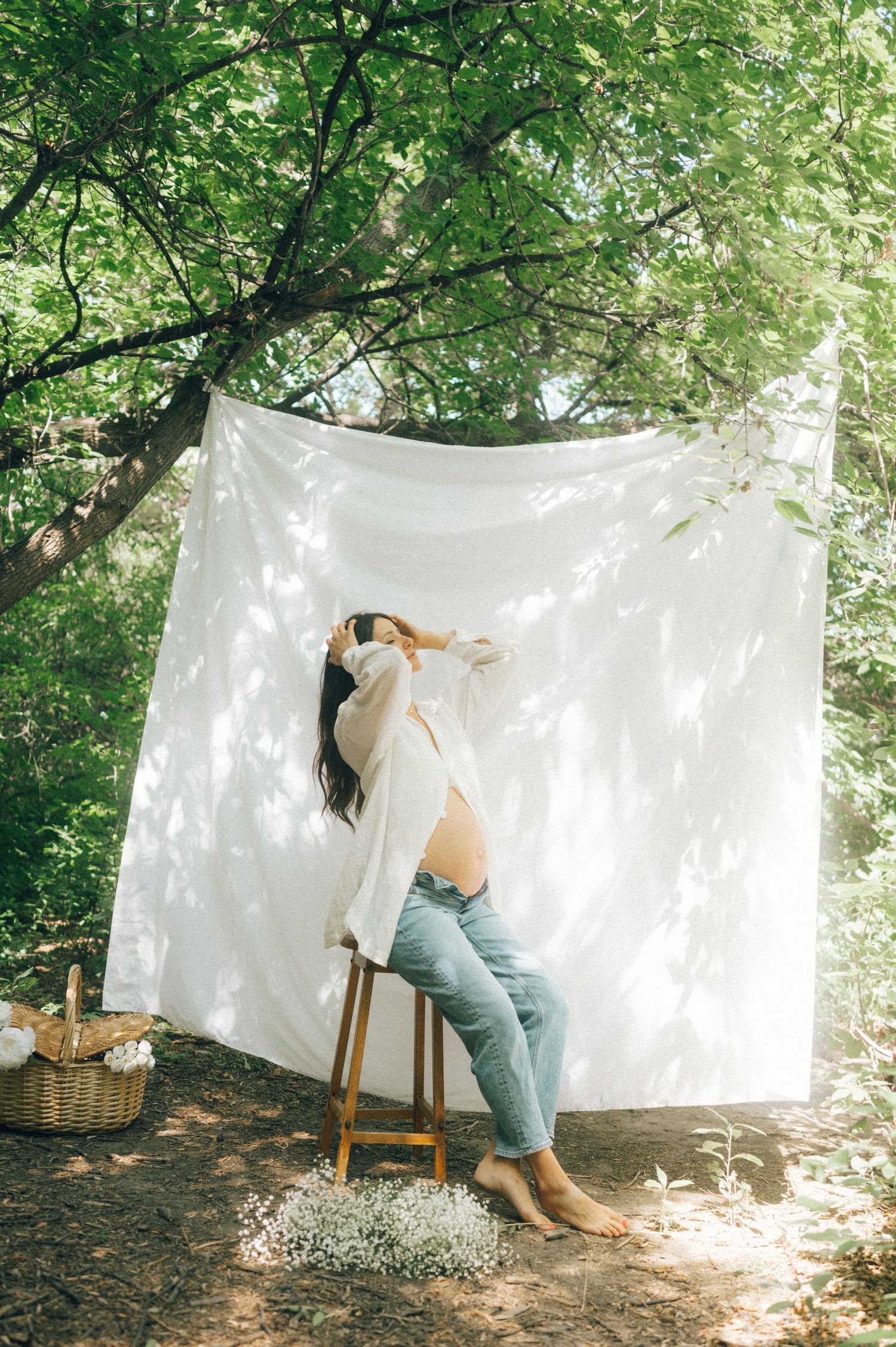 A dreamy outdoor photo shoot setup with a white backdrop sheet hanging between trees and a wooden chair.