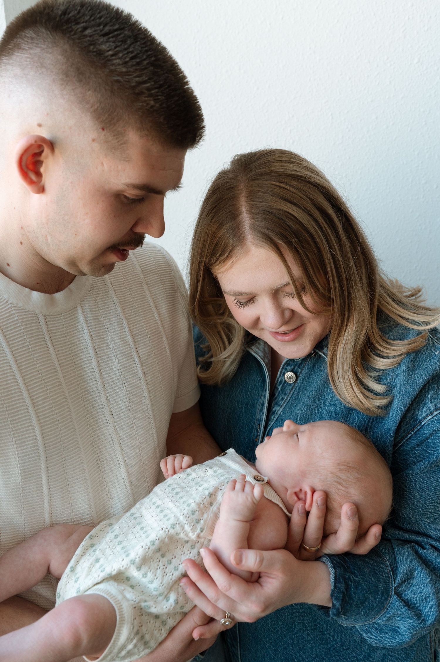 A couple shares a tender moment with their newborn baby, dressed in a white onesie, while sitting together indoors.