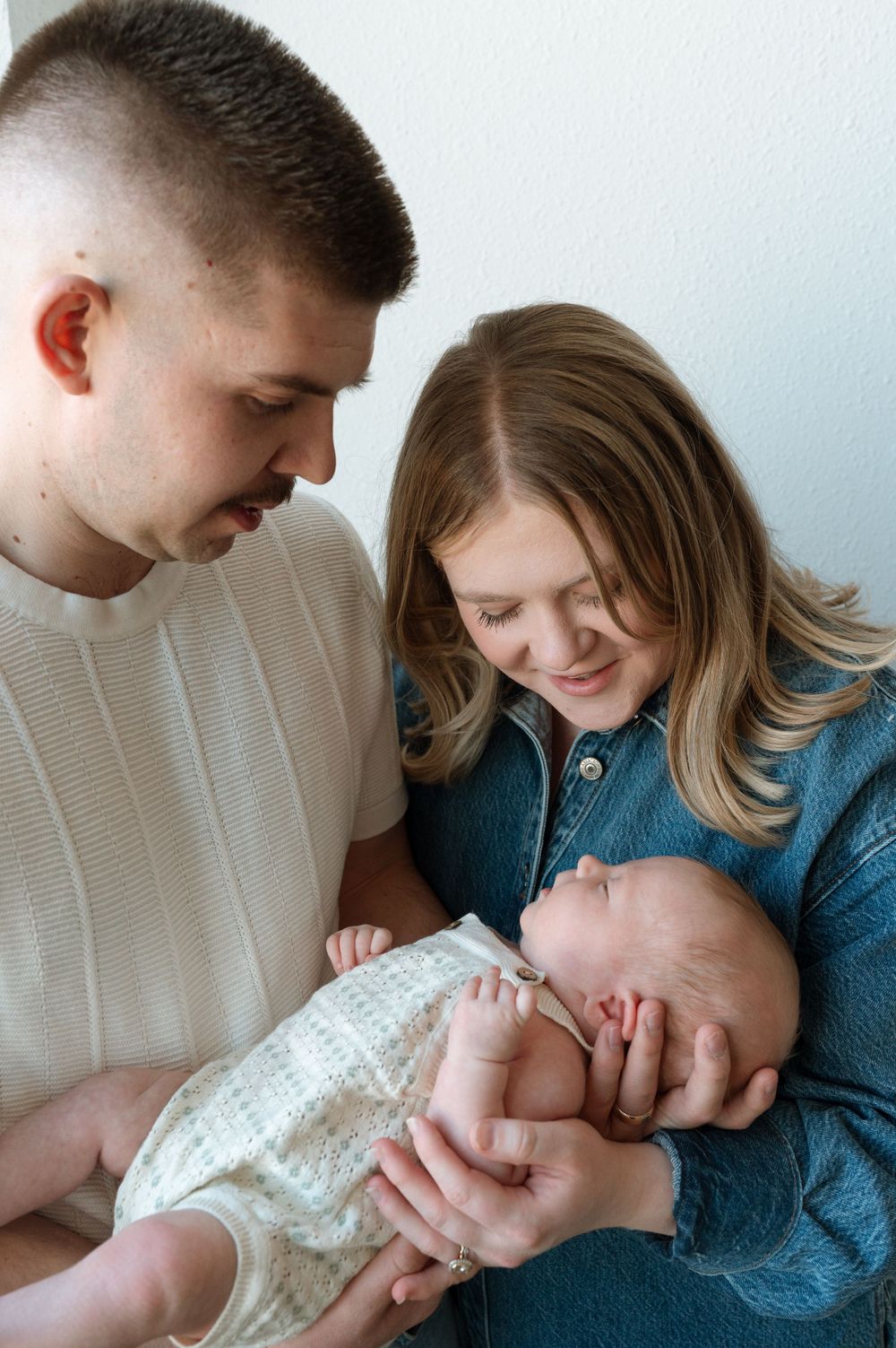 A couple shares a tender moment with their newborn baby, dressed in a white onesie, while sitting together indoors.