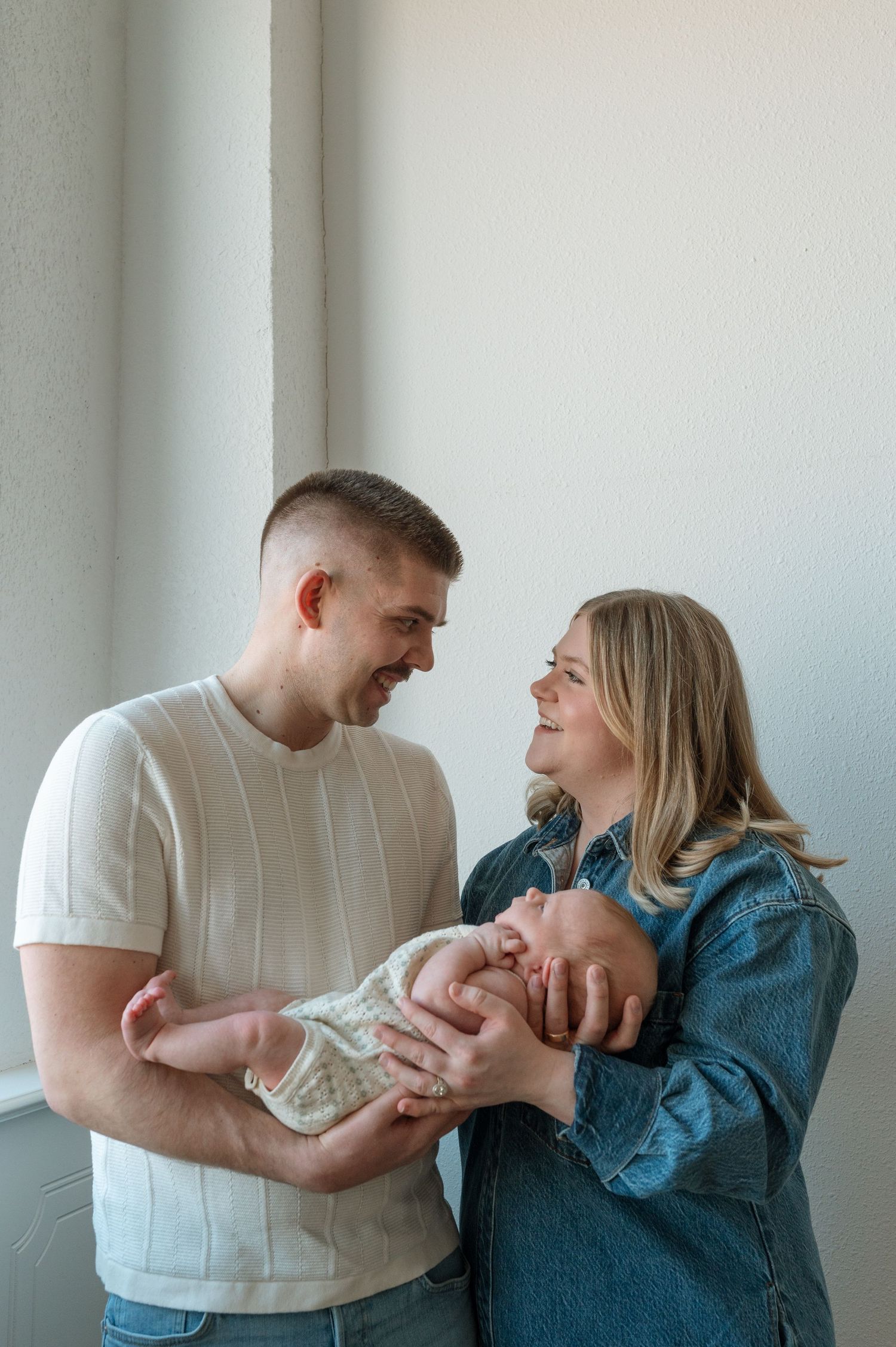 A couple standing by a window lovingly gaze at their newborn baby wrapped in a soft blanket.