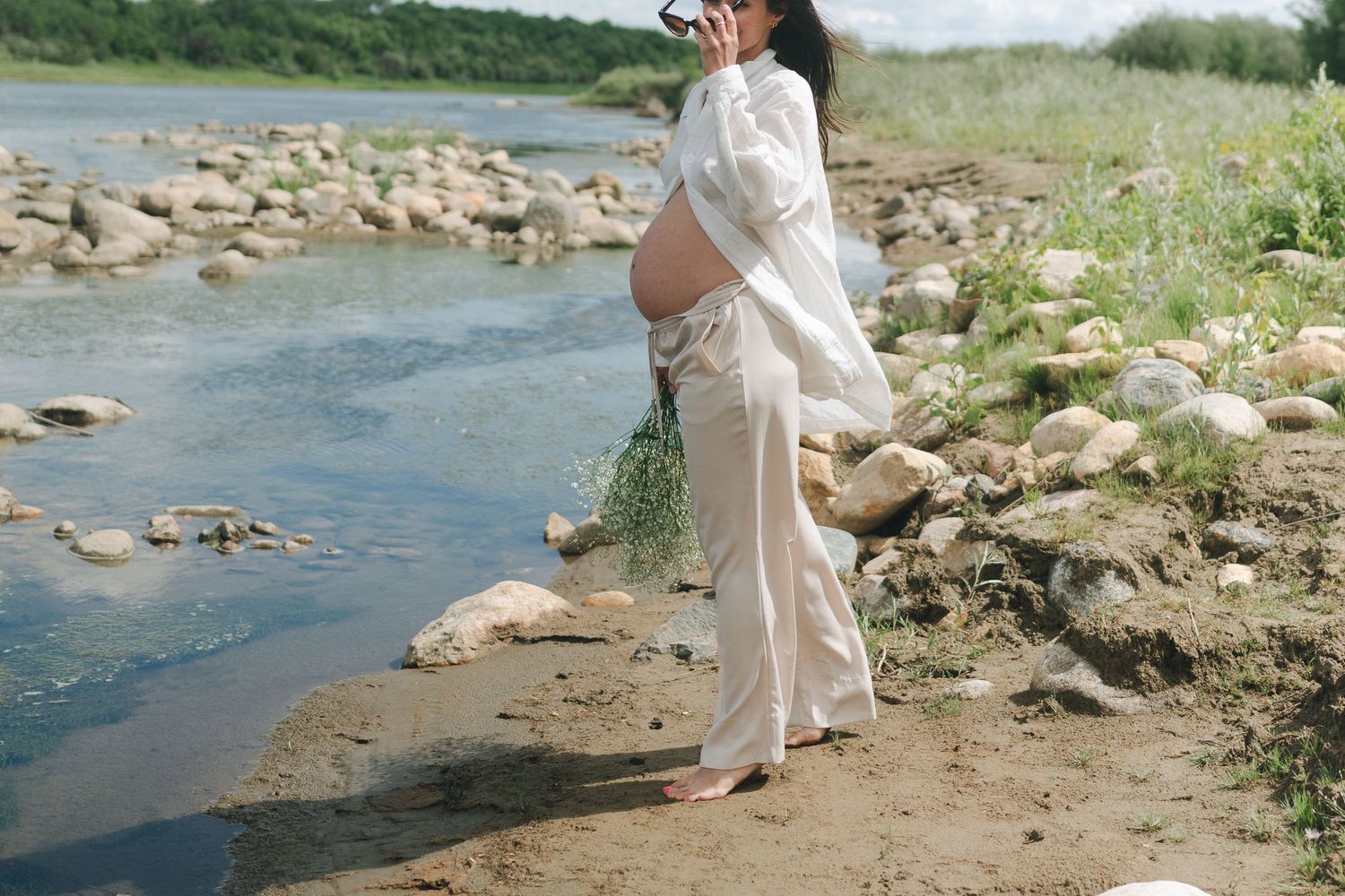 Maternity photo by riverside with flowing white dress and natural scenery.