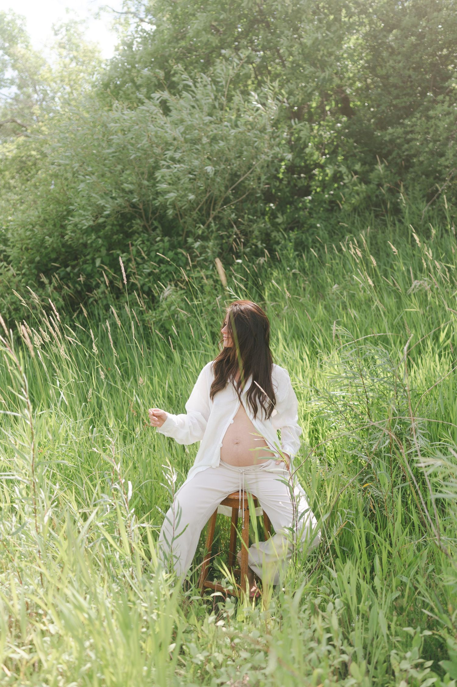 Ethereal maternity portrait in tall grass wearing white clothing during golden hour.