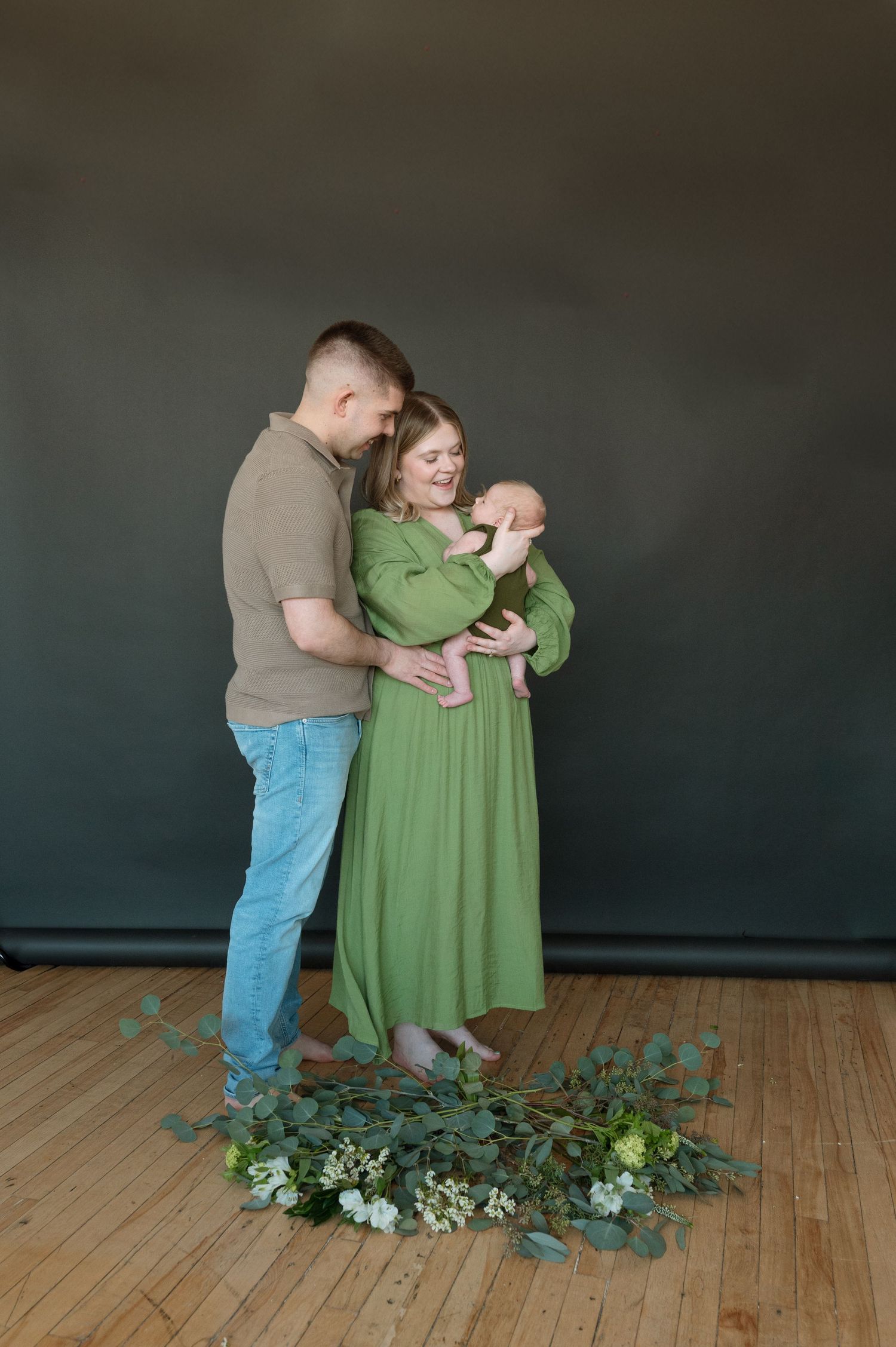 A loving family portrait with parents holding a newborn baby surrounded by floral arrangements on a wooden floor.