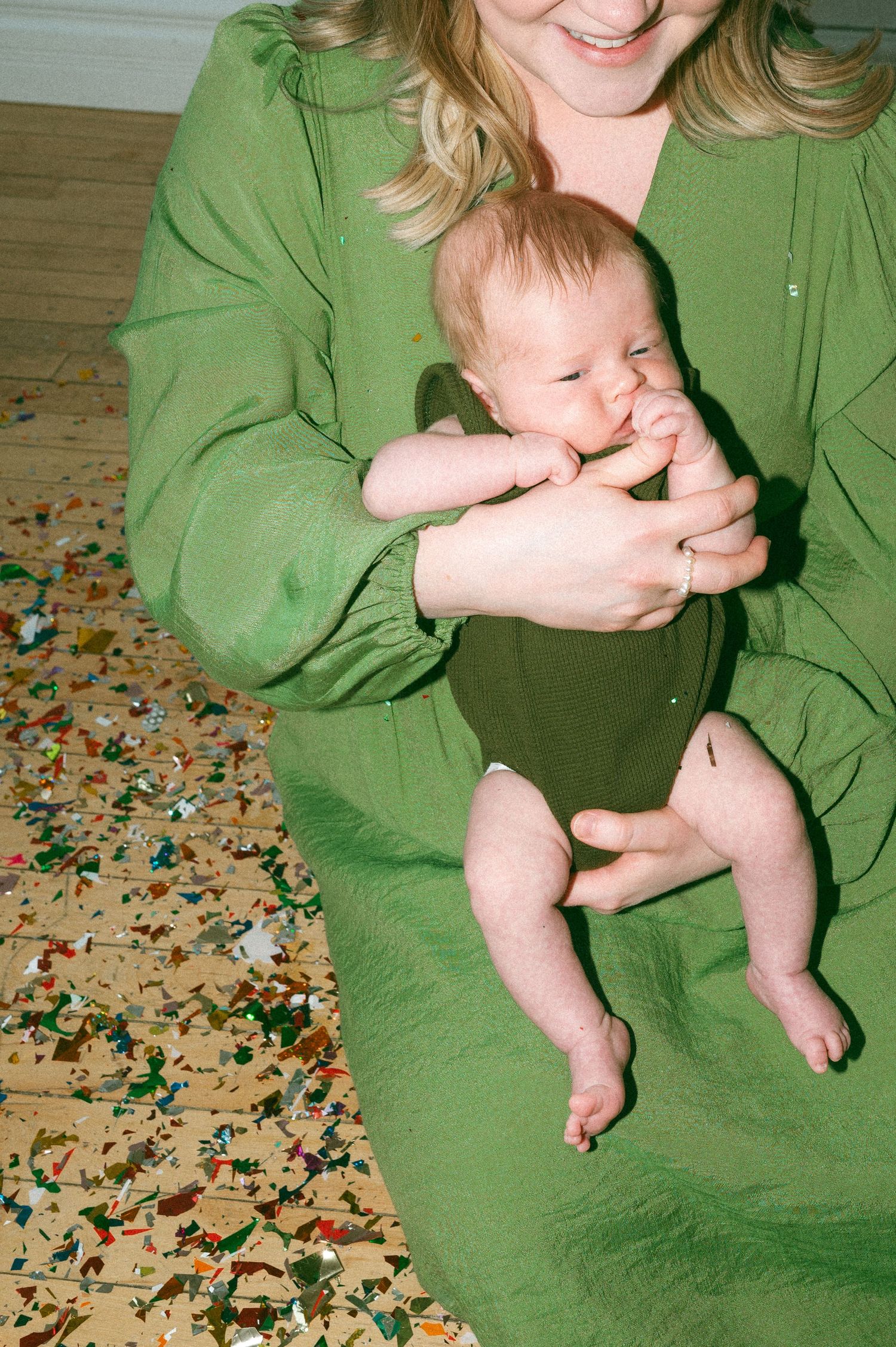 A baby in a green onesie rests on someone wearing a matching green sweater while sitting on a confetti-covered floor.