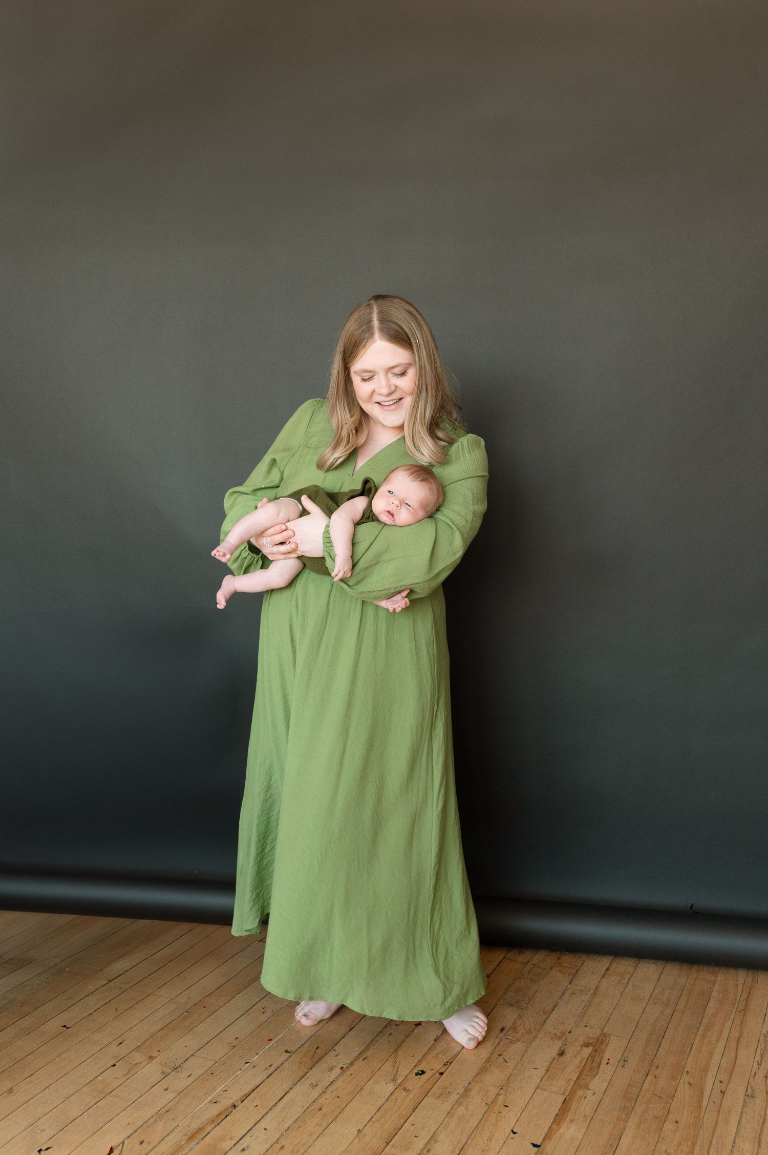 A tender moment captured in a studio portrait as a woman in a flowing sage green dress holds a sleeping baby against dark backdrop.