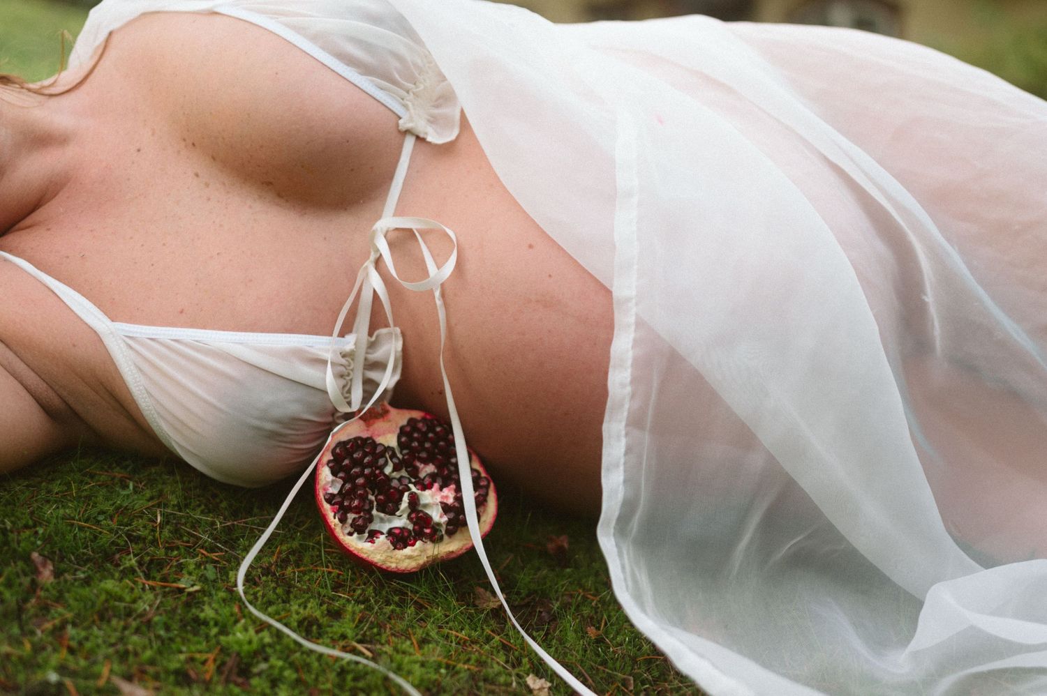 Artistic boudoir photo with flowing white fabric and pomegranate on grass.