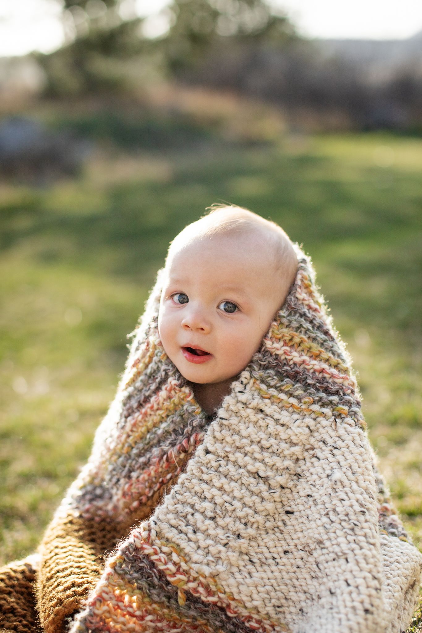 A baby wrapped in a cozy knit blanket smiles while sitting outdoors in warm sunlight.