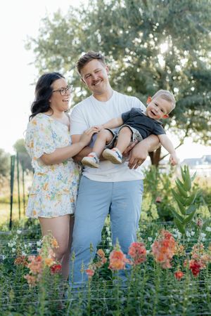 Happy family with parents and baby smiling in a sunny garden with colorful flowers and lush greenery.