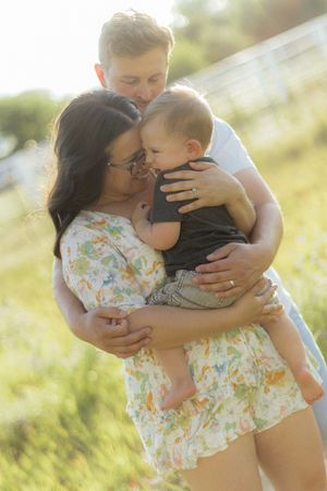 Young couple sharing a tender moment kissing their baby outdoors in a sunlit green field.