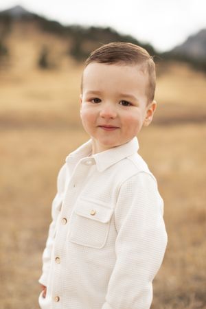 Young child in white button-up shirt poses outdoors with mountainous background.