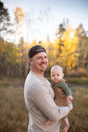 A candid outdoor portrait of a parent holding a baby against an autumn forest backdrop.