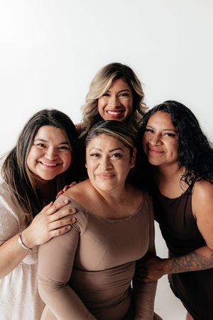 Four smiling women pose together in a warm studio photo, wearing neutral tones against a white background.