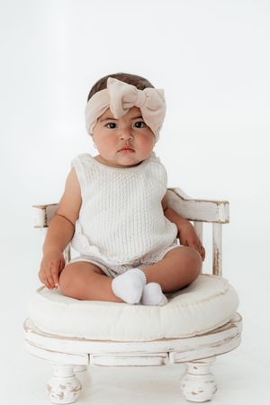 Baby girl in white knit romper and beige bow headband sitting on a vintage white wooden chair.