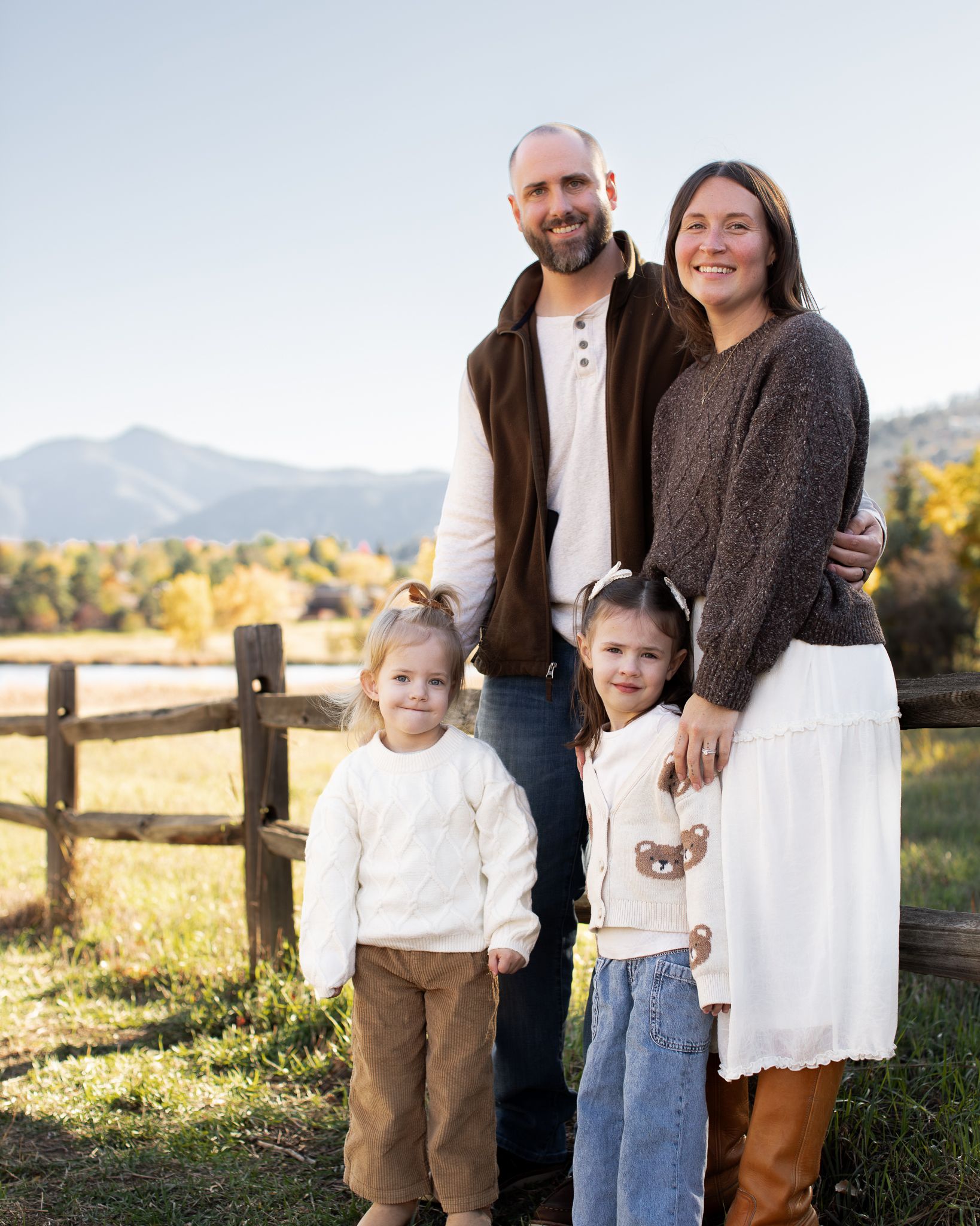 A family poses together outdoors against a rustic wooden fence with mountains and autumn foliage in the background.