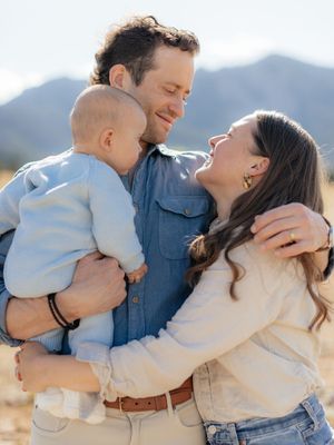 A happy young family with a baby shares a joyful moment outdoors with mountains in the background.