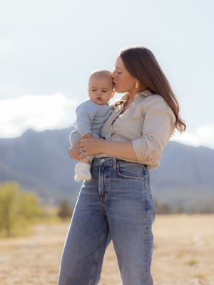 A mother tenderly kisses her baby outdoors with mountain scenery backdrop, both dressed casually in a sunlit field.
