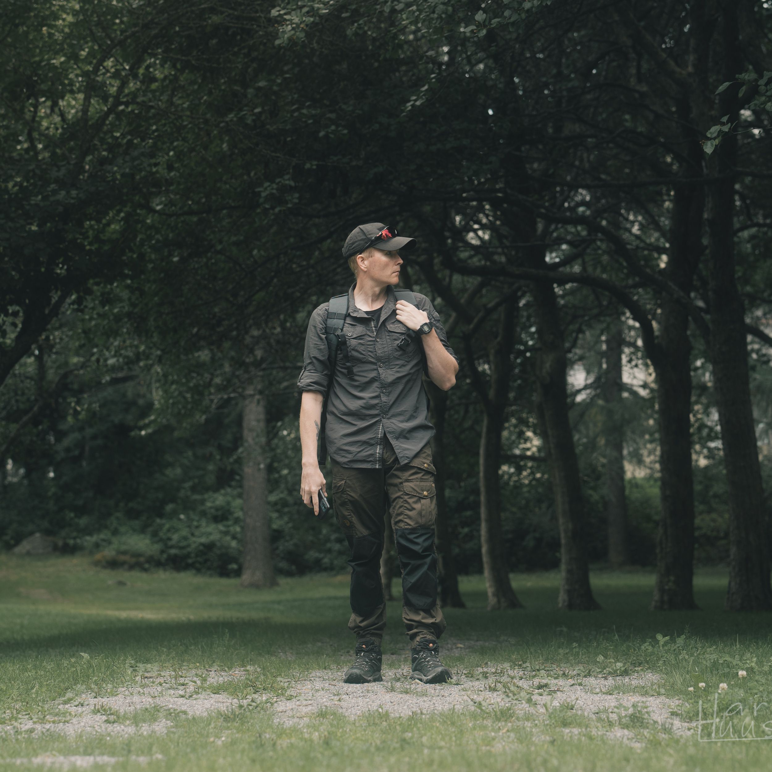 A lone person in casual attire stands on a dirt path in a wooded area surrounded by green trees.